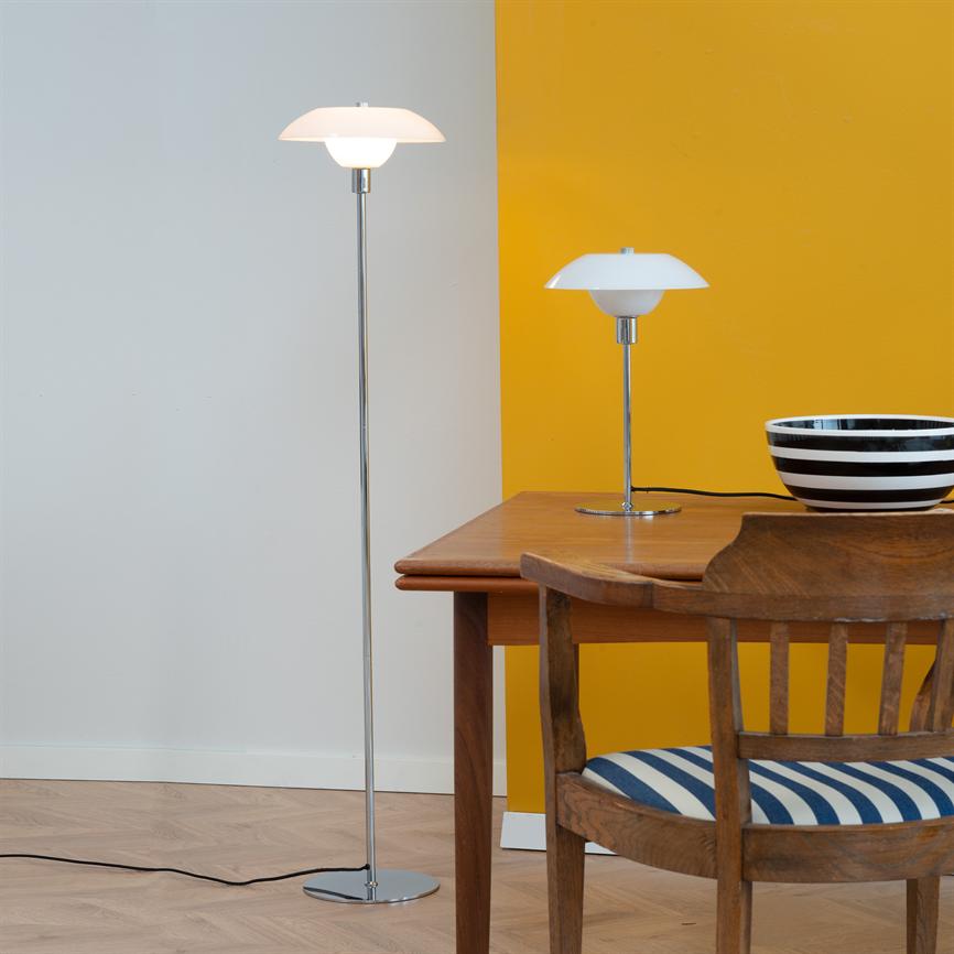 Gulvlampe og bordlampe lyser ved et træbord; en sort-hvid stribet skål står på bordet, og en træstol med blå-hvid stribet pude står ved bordkanten foran en gul og hvid væg.