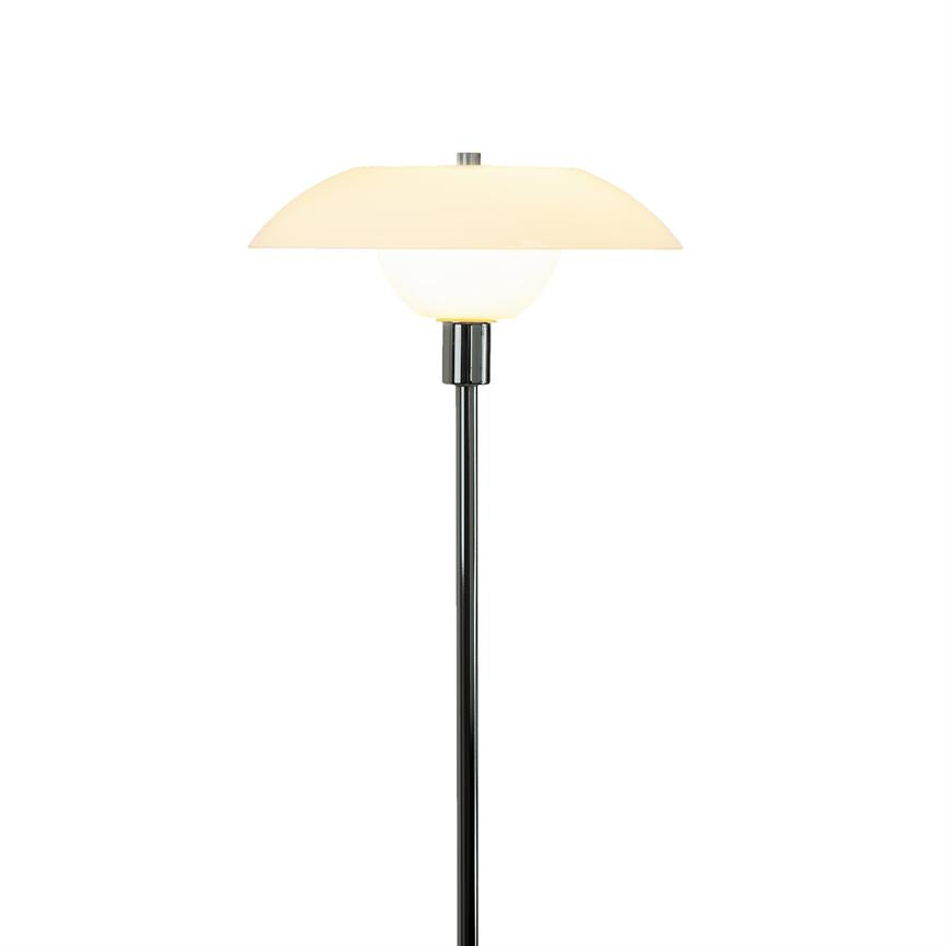 Standerlampe med rund, to-delt opalglasskærm, udsender blødt, varmt lys; står på en slank sort metalstang mod en enkel, hvid baggrund.