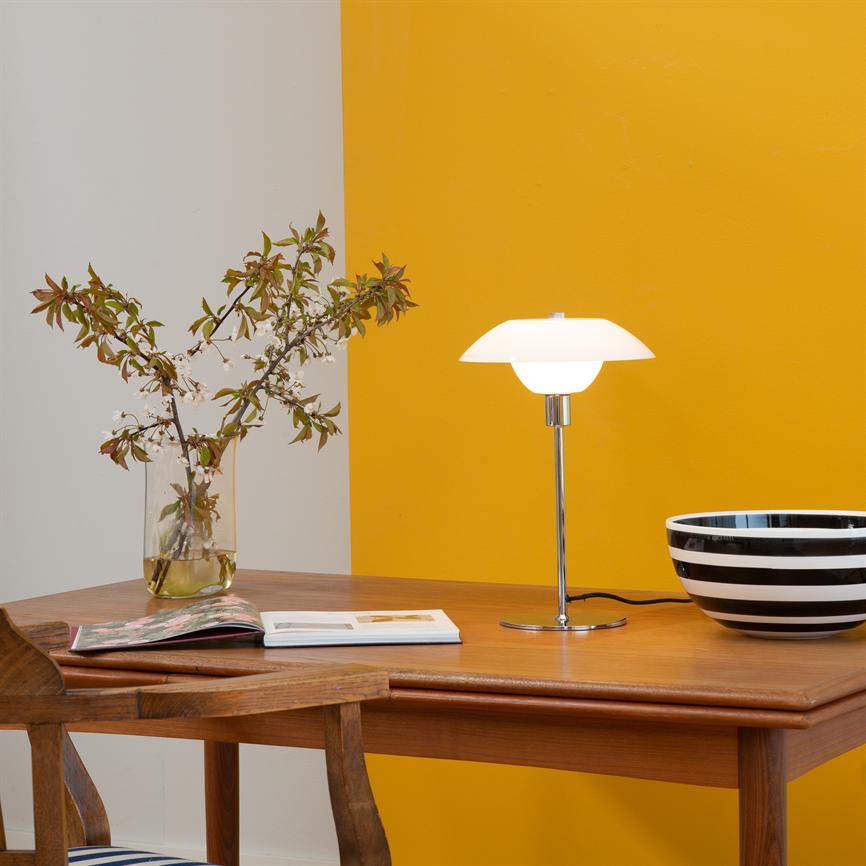 Bordlampe lyser blødt over et træbord; til venstre glasvase med blomstergrene og åben bog, til højre sort‑hvid stribet skål; væg i baggrunden delt i hvid og gul.