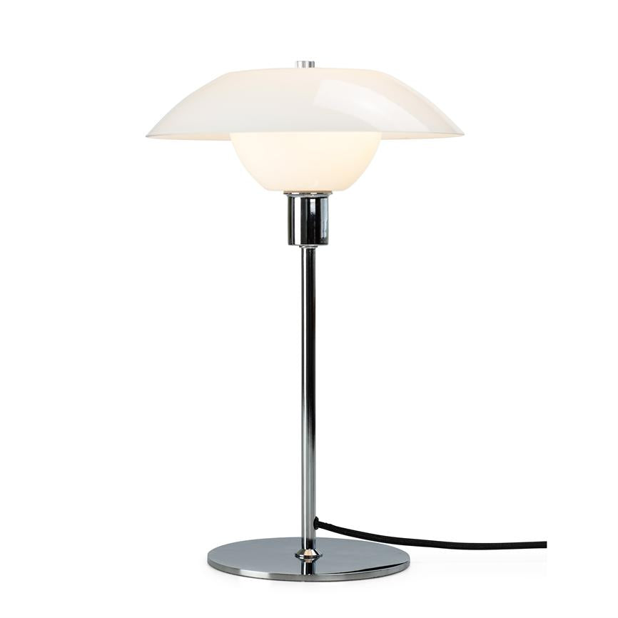 En bordlampe står tændt og afgiver blødt, varmt lys gennem en rund hvid skærm; den er monteret på en slank kromstang med rund kromfod og sort ledning på hvid baggrund.