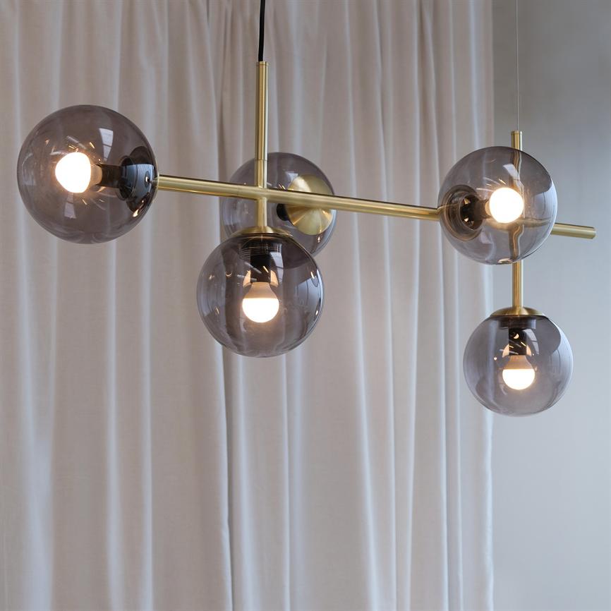 En moderne loftslampe hænger med fem røgfarvede glaskugler, nogle med tændte pærer; en messingstang forbinder kuglerne — foran bløde hvide gardiner i et lyst rum.