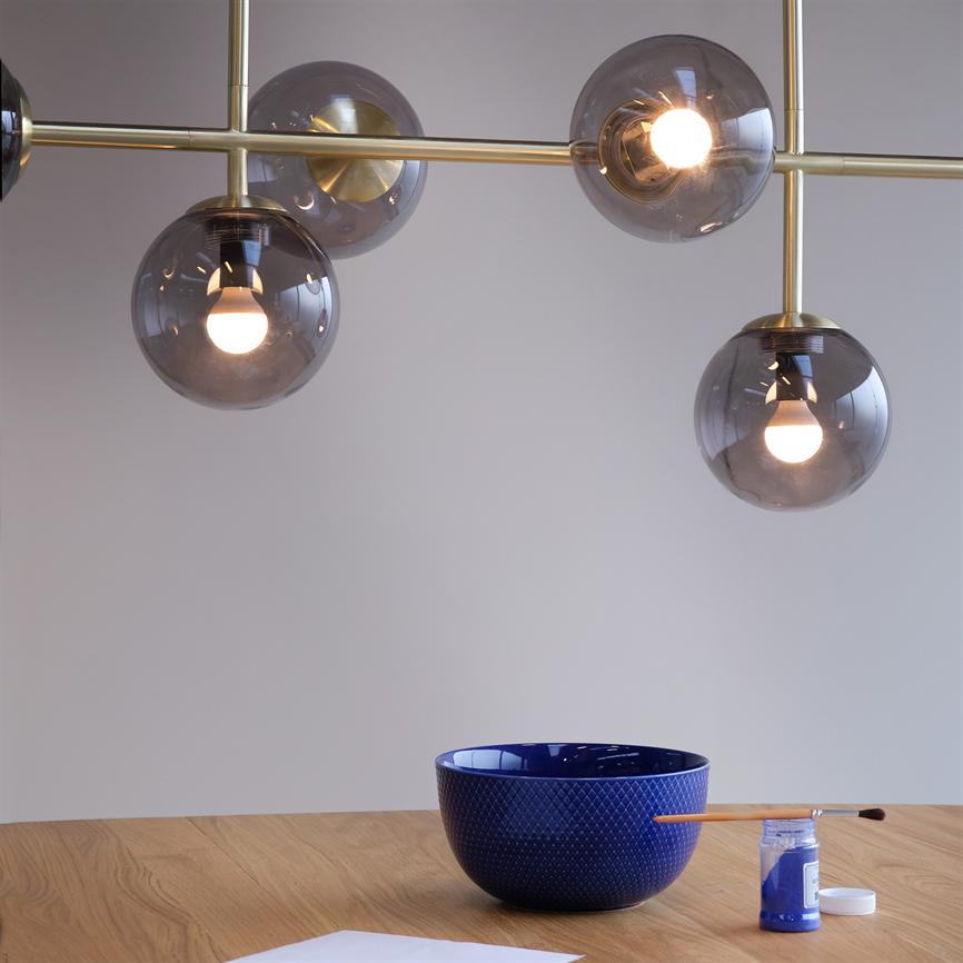 Moderne loftslampe med røgfarvede glaskupler, hvor tre pærer lyser, hænger over et træbord med en mørkeblå skål, en pensel og en åben krukke blå maling.