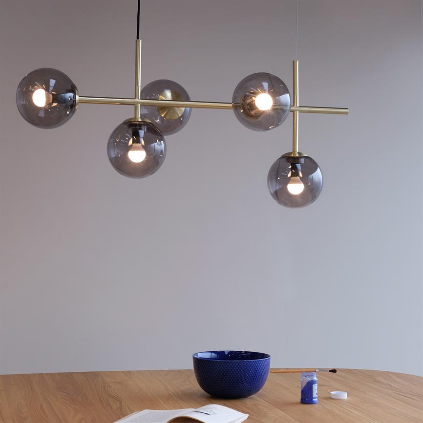 Moderne loftlampe med fem røgfarvede glaskupler hænger og spreder varmt lys over et træbord; et minimalistisk rum med en blå skål, åben bog og en lille bøtte maling.