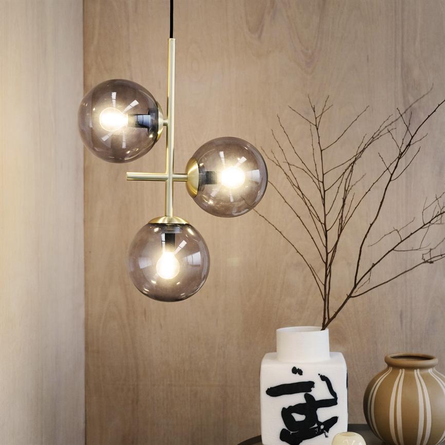 Ophængt lampe med tre røgfarvede glaskupler udsender varmt lys foran lyst træpanel ved et bord med hvid vase med sort penselstrøg, beige stribet vase og tør gren.