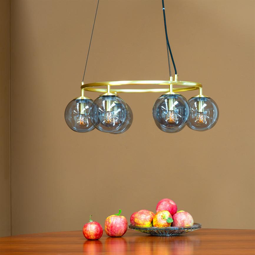 En gylden rund loftslampe med fire røgfarvede glasglober hænger over et træbord, med lysende filamenter; en skål med røde æbler står mod en beige væg.