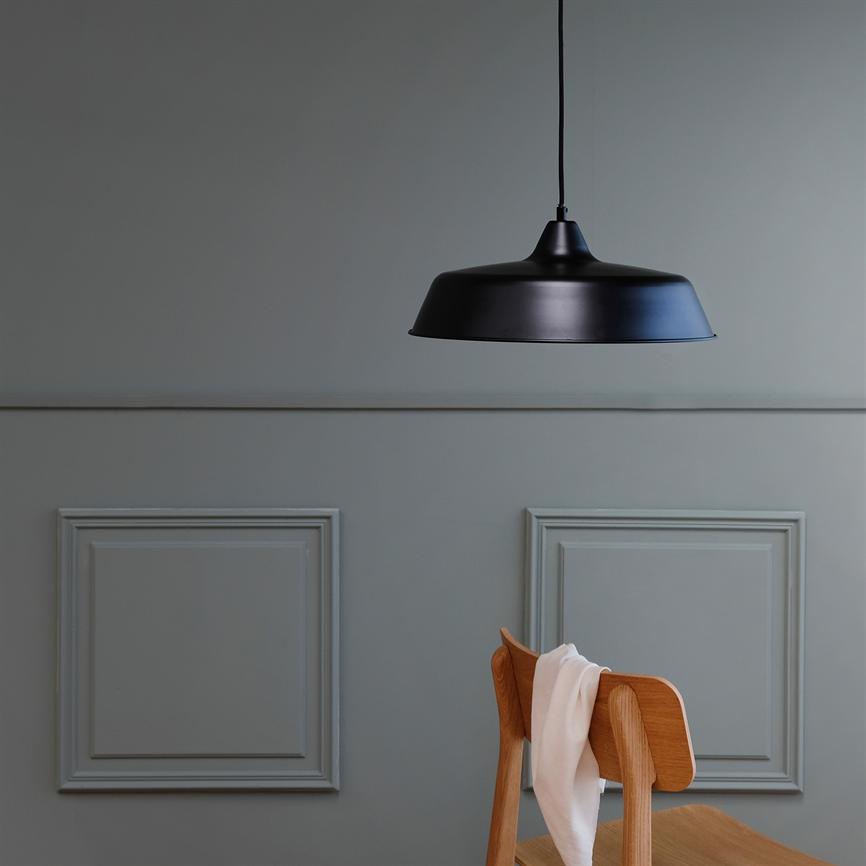 Sort metal loftlampe hænger over en træstol, hvor et hvidt stykke stof er kastet; omgivet af en dæmpet grå væg med to dekorative panelrammer.