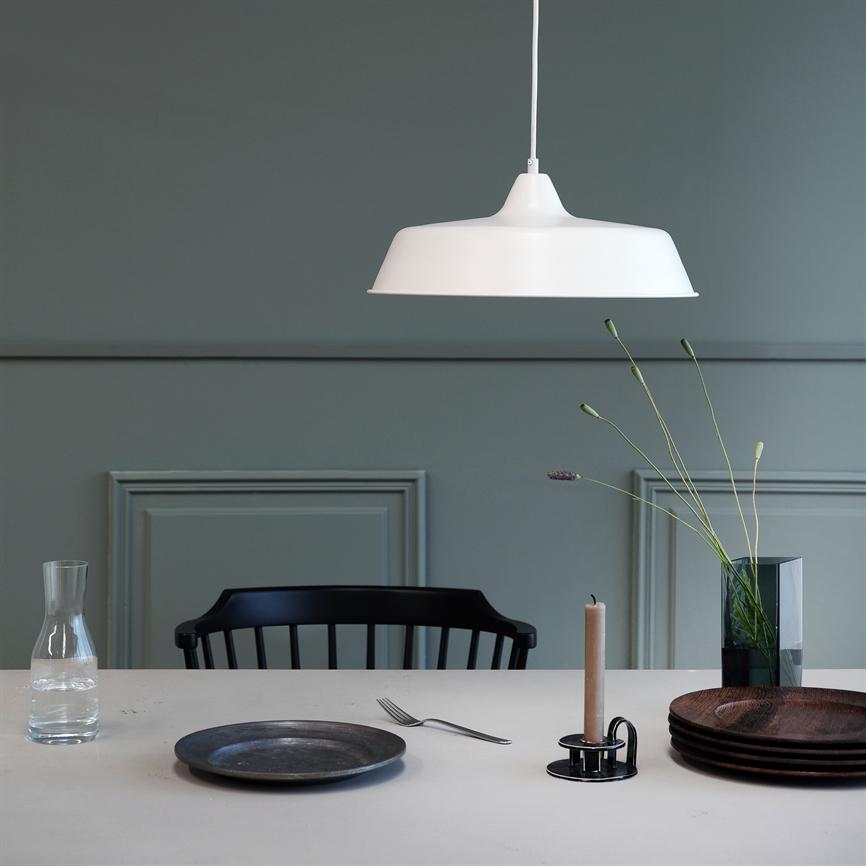 Hvid pendel hænger over et dækket bord; en sort stol bagved, glas-karaf, en tallerken og gaffel, lysestage med lys og vase med høje blomster foran grøn væg.