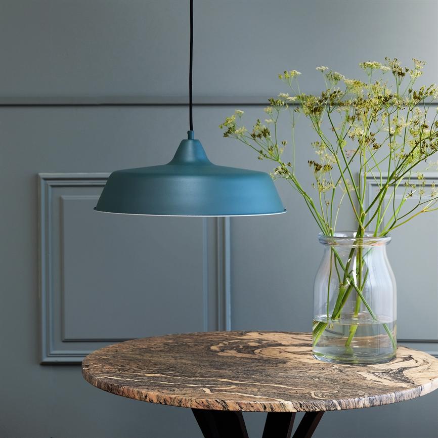 En blå pendel hænger over et rundt marmorbord; en glasvase med sarte hvide blomster står på bordet foran en gråblå væg med indrammede paneler i et roligt, minimalistisk interiør.