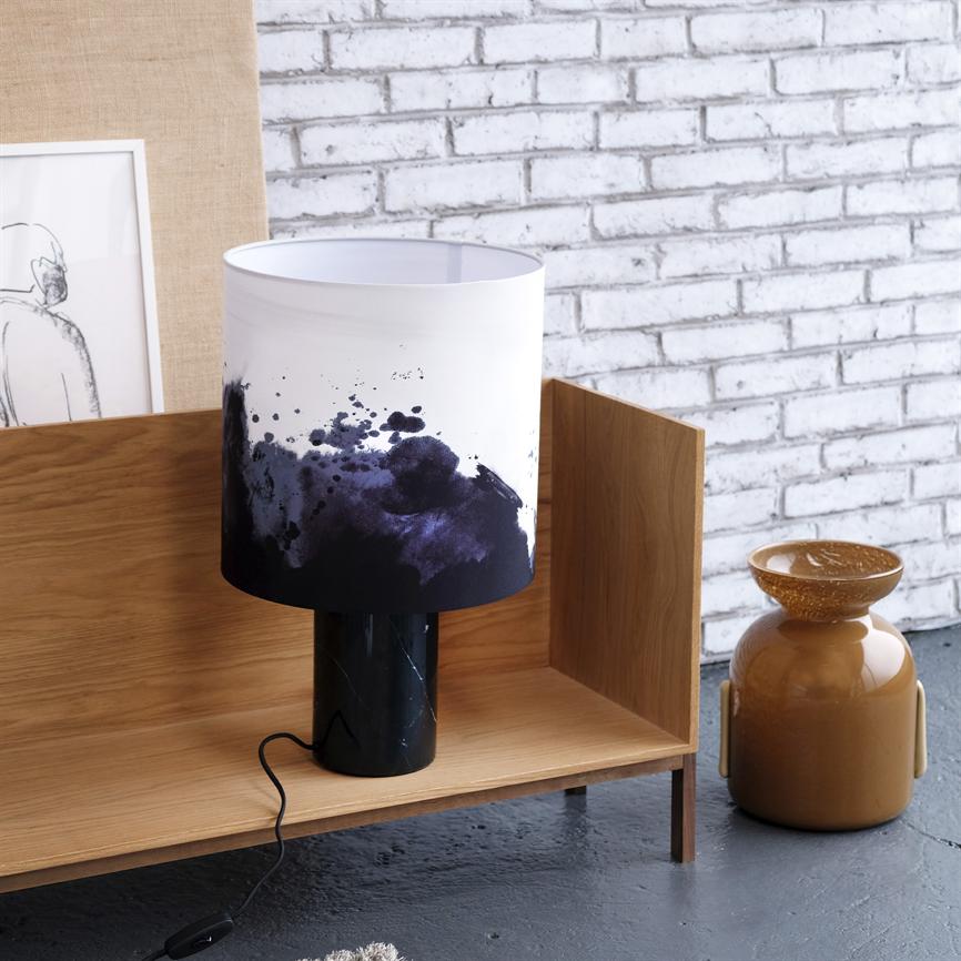 En bordlampe med hvid skærm plettet af mørkeblå akvarelstænk står på en lav træhylde; sort marmorfod og ledning, foran hvid murstensvæg, med en brun keramisk vase og indrammet skitse.