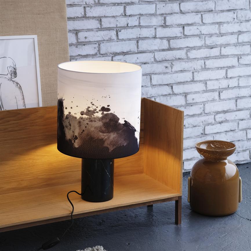 En bordlampe står tændt på en lav træhylde, kaster varmt lys mod en hvid murstensvæg; til højre en brun keramikkrukke, til venstre en indrammet skitse.
