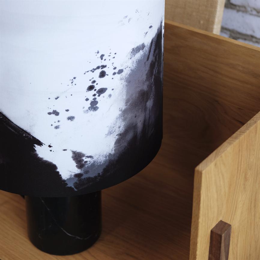 Lampe med sort-hvidt pletemønstret skærm står på en sort marmorfod inde i en lys træbakke på træbord, tæt ved en rå murstensvæg.