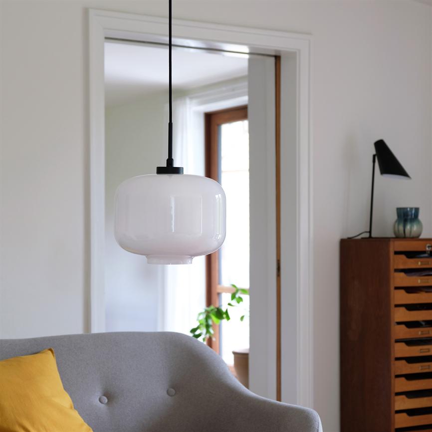 En hvid, rund glaspendel hænger i midten; i forgrunden en grå sofa med gul pude, i baggrunden en trækommode med bordlampe, døråbning og en grøn plante ved vinduet.