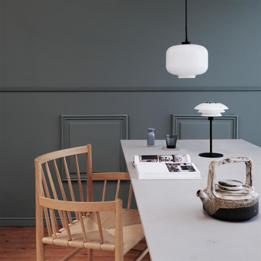 En hvid pendel lyser over et minimalistisk bord; en åben bog og en keramisk tepotte ligger på bordet, en træstol trukket til, mørkegrå vægge og trægulv i baggrunden.