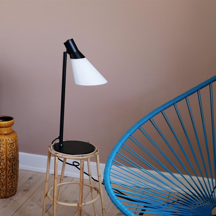 Bordlampe med sort skaft og hvid skærm peger diagonalt nedad fra en bambus skammel; ved en rosa væg på lyst trægulv, flankeret af en blå snorestol og en gul dekorationsvase.