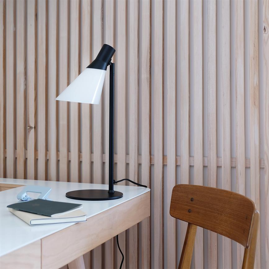 Sort-hvid bordlampe står på et lyst skrivebord ved en træstol, foran en væg med lodrette lyse trælameller; en notesbog og små skrivegenstande ligger på bordet.