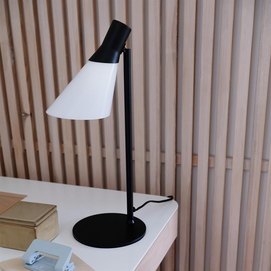 Skrivebordslampe står med skråt nedadrettet hvid skærm på sort stang, placeret på hvidt bord foran lodrette trælameller; guldæske og lyseblåt hulapparat ved siden af.