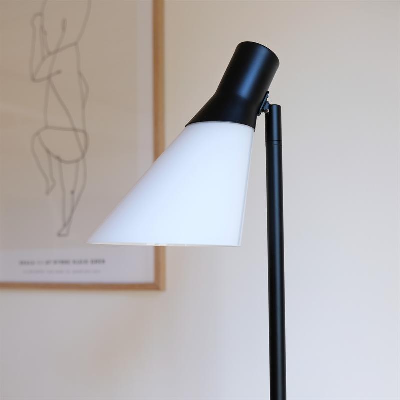 Gulvlampe med hvid, kegleformet skærm peger nedad; sort stang og fatning. Baggrunden viser en sløret stregtegning på en lys væg i et minimalistisk rum.