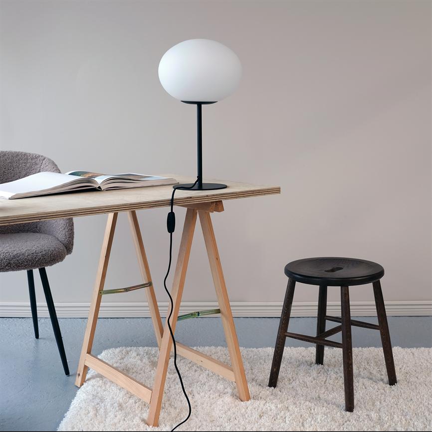 En rund bordlampe står på et træbord ved en åben bog, omgivet af en polstret stol, en mørk skammel og et lyst, minimalistisk rum.