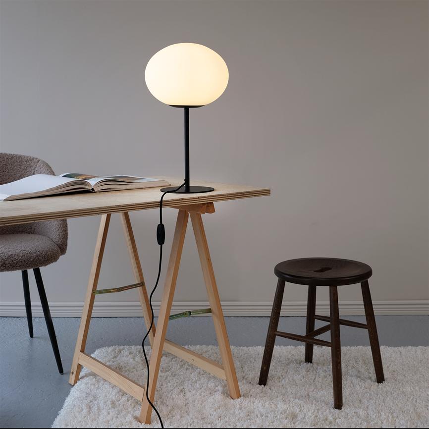 Bordlampe lyser blødt og står på et enkelt træbord med savben; en åben bog, en stol og en mørk træskammel ved siden af i et roligt, minimalistisk rum med lyst tæppe.
