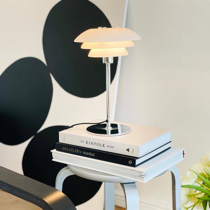 En moderne bordlampe står tændt på en stak bøger på en lille hvid skammel; sorte grafiske cirkler på væggen bagved og en blomst i nederste højre hjørne. 
 Tekst i billedet: THE KINFOLK HOME; ARA GÜLER's İstanbul