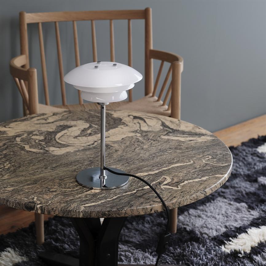 Bordlampe står på et rundt marmorbord med beige og mørke årer; sort ledning løber ud over kanten. Bagved en træstol mod grå væg og et mørkt, blødt tæppe.
