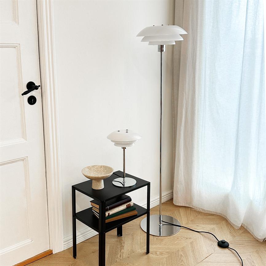 Hvid, lagdelt gulvlampe står ved et lyst vindue; en matchende bordlampe står på et lille sort sidebord med bøger og skål på lyst trægulv ved en hvid dør.
