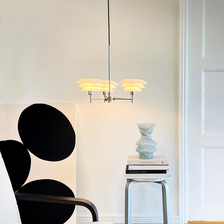 Loftlampe hænger oplyst over et sidebord med stablede bøger ('THE KINFOLK HOME'; 'ARA GULER Istanbul') og en snoet glasvase, ved en hvid væg med sort-ovalt maleri og lænestol.