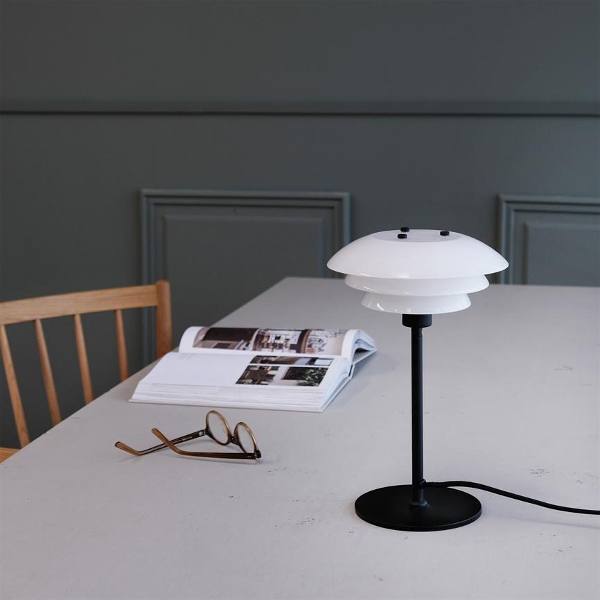 Hvid, skålformet bordlampe står på et gråt bord; ved siden af ligger briller og et åbent magasin; rummet har en grå væg og en enkel træstol.