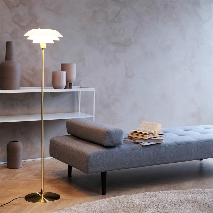 Gulvlampe lyser blødt over en grå dagsseng, hvor en stak åbne og lukkede bøger ligger; minimalistisk stue med lyse pudrede vægge, hvid hylde og keramiske vaser.