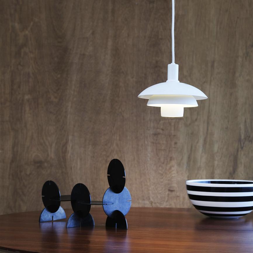 En hvid pendel lampe hænger og kaster blødt lys nedover et træbord; på bordet står tre sorte cirkulære skulpturer og en sort-hvid stribet skål foran en varm træbeklædt væg.