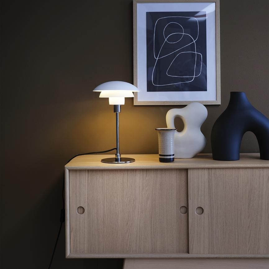 En hvid bordlampe står tændt på en lys egetræsskænk; den kaster varmt lys over en keramikkrukke og to skulpturelle vaser under en indrammet, sort-hvid abstrakt plakat på en brun væg.