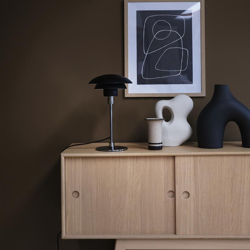 En lys egetræs-skænk bærer en sort bordlampe, et keramikkrus og to skulpturelle vaser foran en brun væg med en indrammet abstrakt sort-hvid plakat.