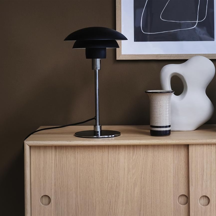 En sort bordlampe står på et lyst egetræsskab; ved siden af står en keramisk vase og en hvid abstrakt skulptur foran en indrammet sort-hvid grafisk plakat på en brun væg.