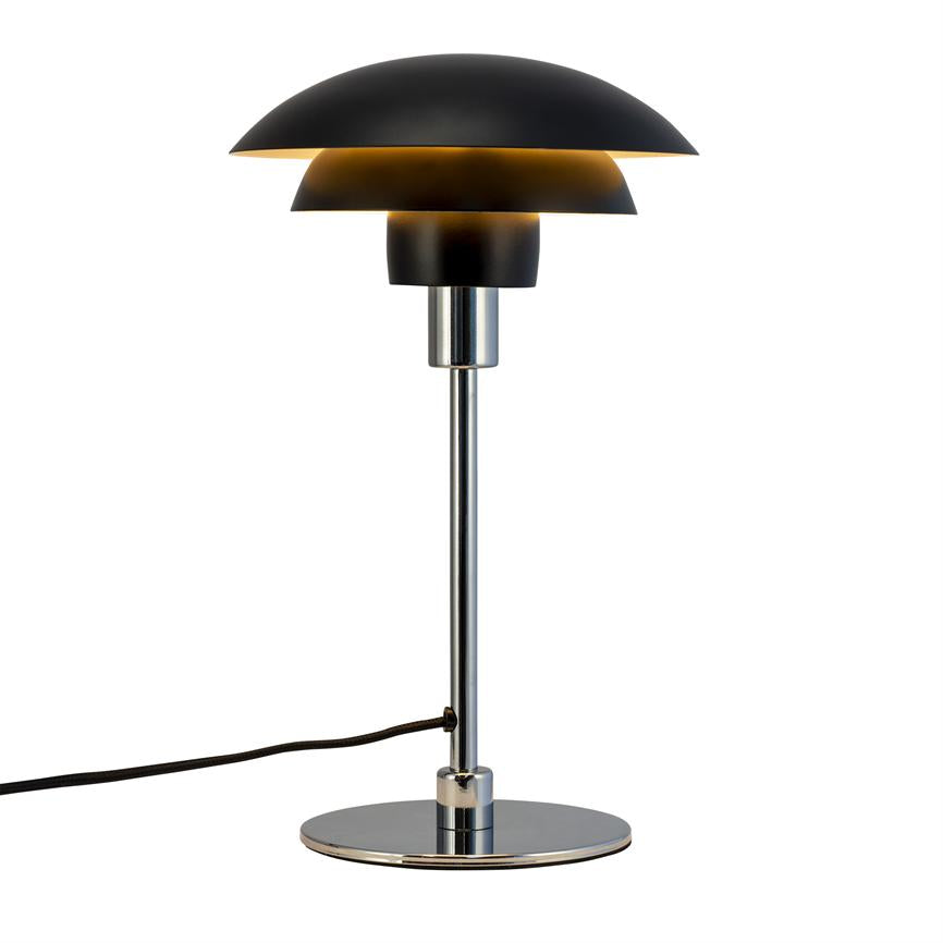Bordlampe med lagdelt sort skærm udstråler varm, gylden glød; slank kromstang og rund fod står mod hvid baggrund, ledning ligger synlig.