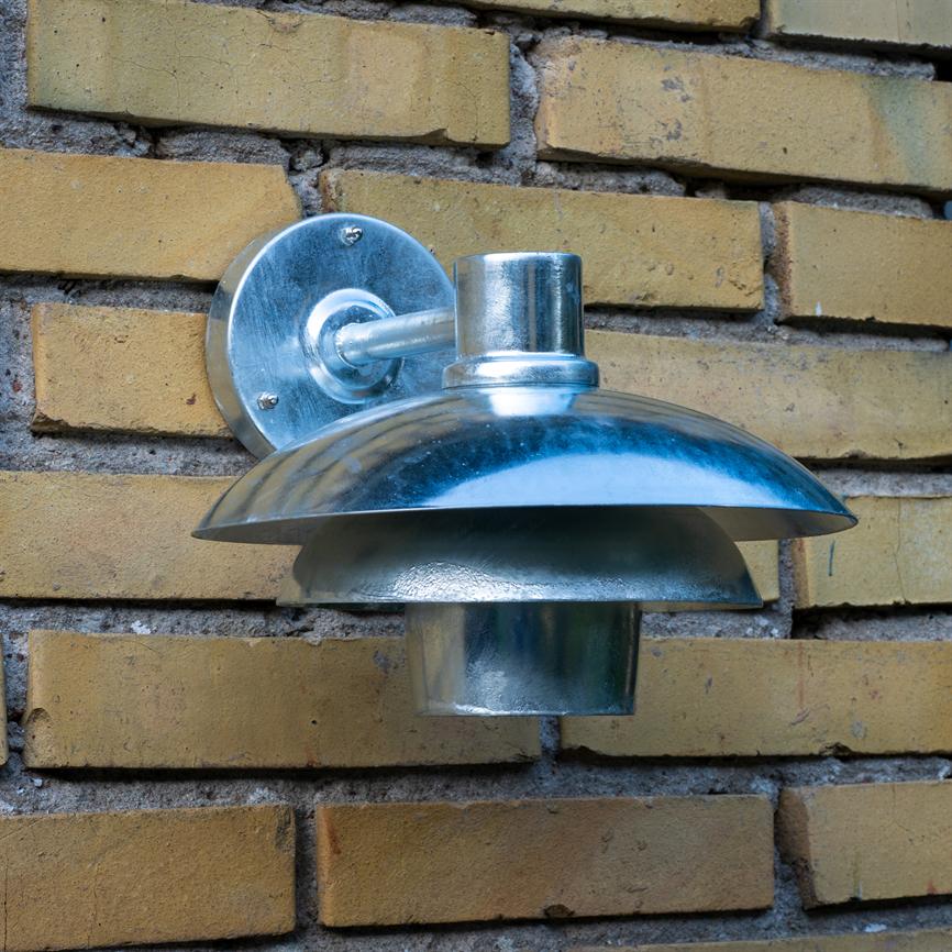 Metal udendørslampe monteret på en gul murstensvæg; rund, blank skærm reflekterer lys, kort arm og rundt vægbeslag fastgør lampen, omgivet af vandret lagte gule teglsten.