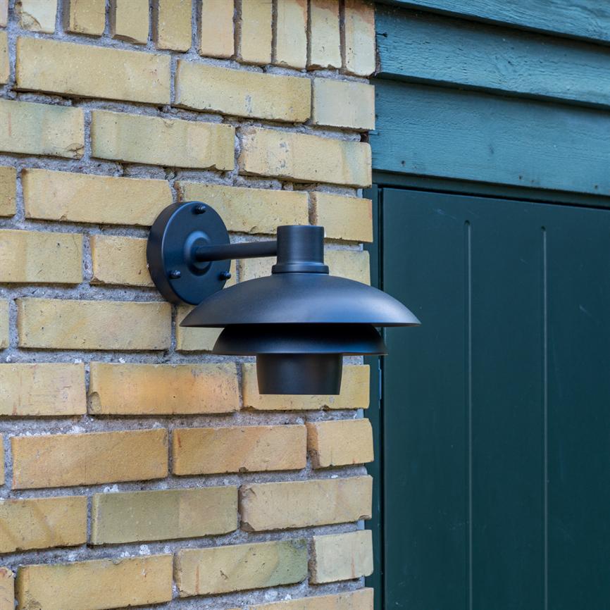 Væglampe i sort metal er monteret på en gul murstensvæg, peger nedad og hænger fra et rundt vægbeslag; den sidder ved siden af en mørkegrøn trædør og træbeklædning.