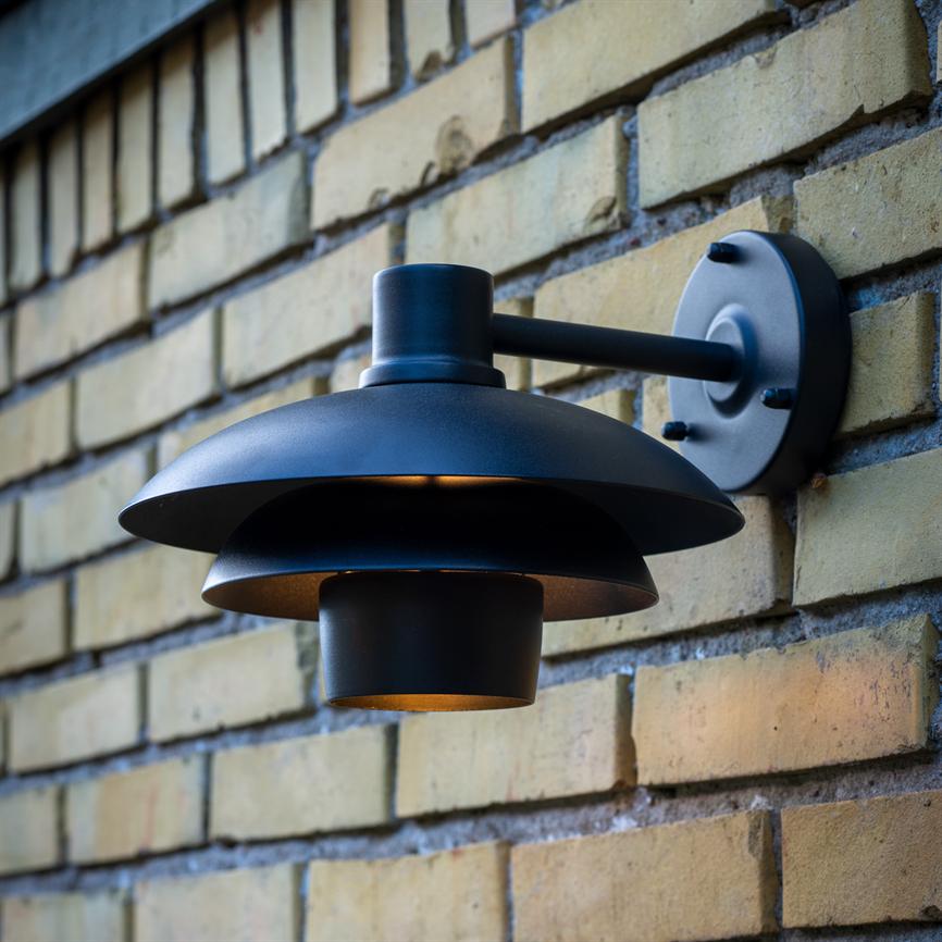 En sort, todelt metal væglampe udsender et varmt, nedadrettet skær; den er monteret på et rundt vægbeslag og sidder på en gulbrun murstensfacade.
