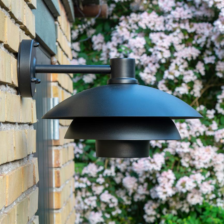 Sort, mat, trelags udendørs væglampe monteret på en gul murstensvæg; en rund arm holder lampen ud fra facaden, med grøn busk dækket af lyserøde blomster i baggrunden.