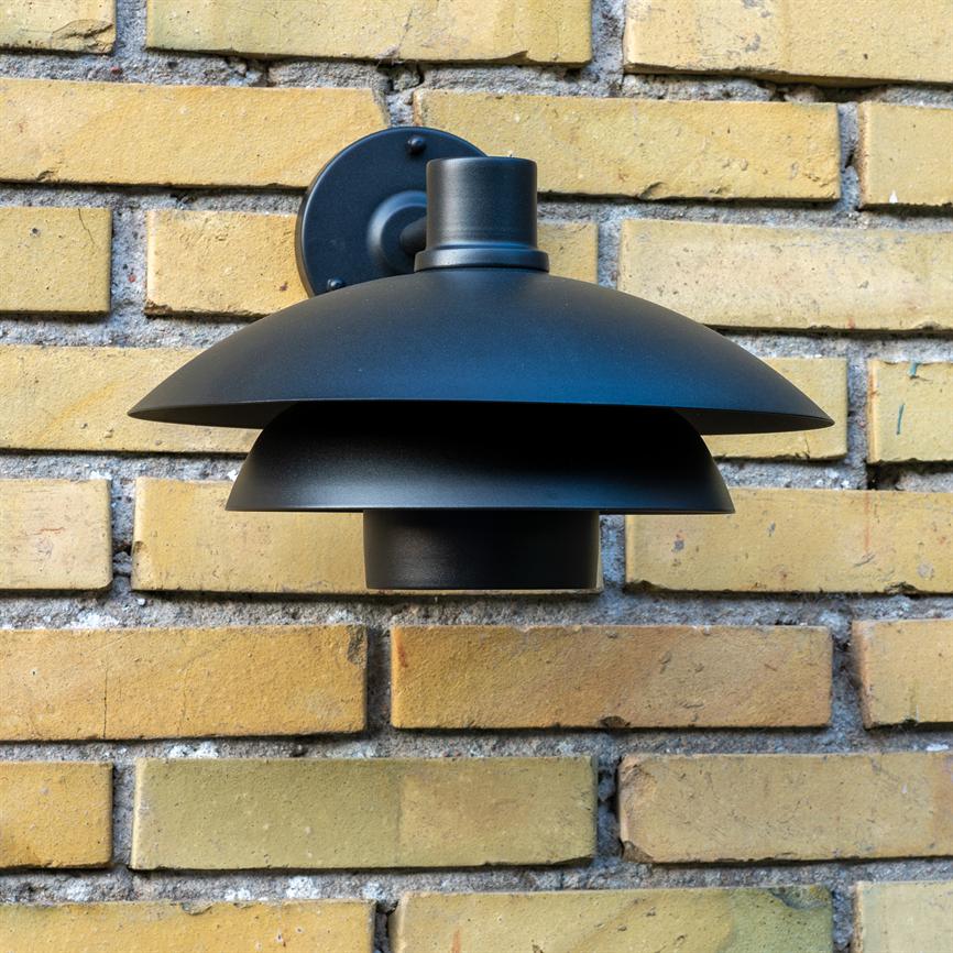 Sort metal væglampe er monteret på en gul murstensvæg; tre brede, nedadrettede skærme danner et lagdelt, moderne udendørsarmatur.