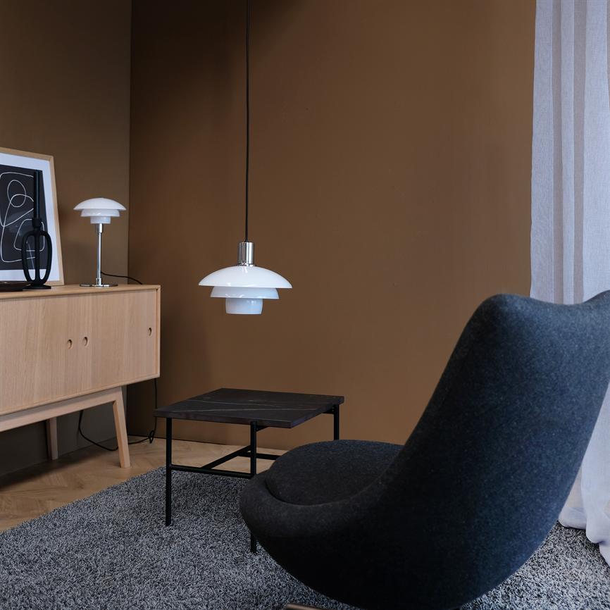 Hvid lagdelt pendellampe hænger over et lille sort sofabord i et roligt hjørne med en mørkegrå lænestol, lyst egetræs-skænk med bordlampe og dekor, gråt tæppe og brun væg.