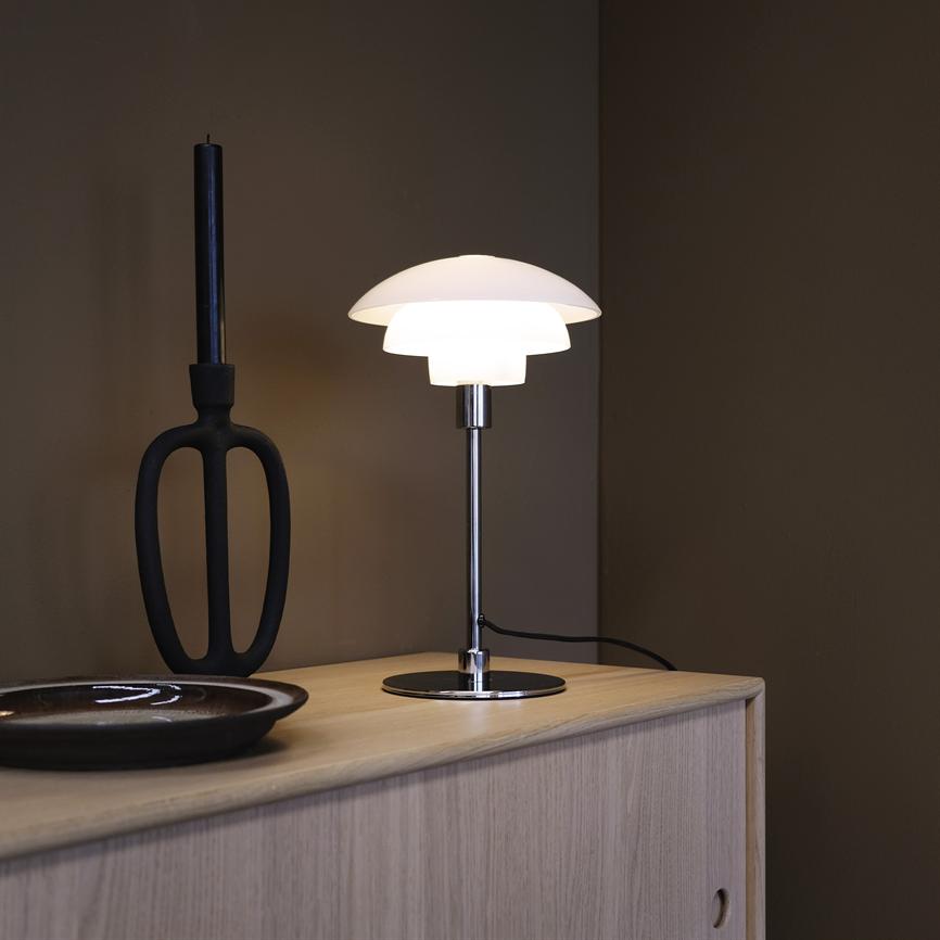 Bordlampe lyser blødt på en lys trækommode foran en mørk væg; ved siden står en sort lysestage og en mørk, rund keramisk skål.