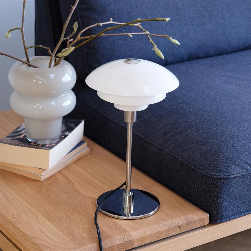 Bordlampe står slukket på et lyst træbord; hvid glasskærm og kromfod, ved siden af en hvid vase med grene og en blå sofa i baggrunden.