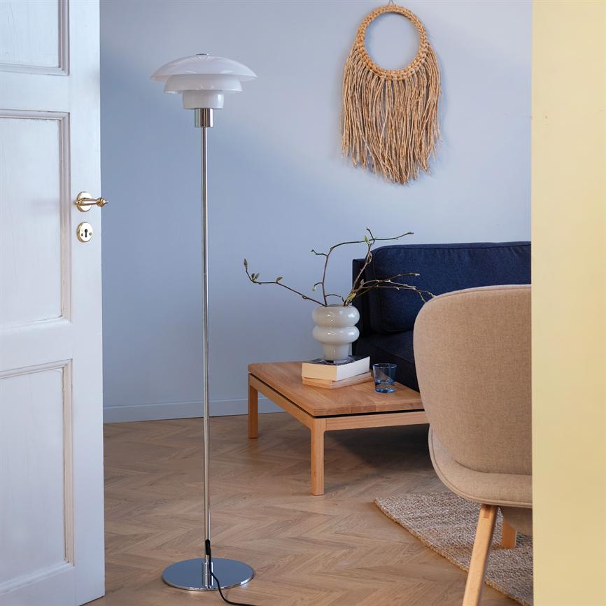 En slank metal gulvlampe står ved et lavt træ-sofabord; omkring ligger en mørk blå sofa, en beige lænestol, en vase med grene og et rundt makramé vægophæng i en lys stue.