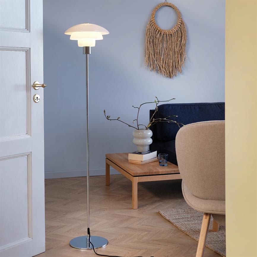 En gulvlampe står tændt ved et lavt sofabord; en keramisk vase med grene, en mørkeblå sofa og en makramé-vægdekoration i et lyst, nordisk stuerum.