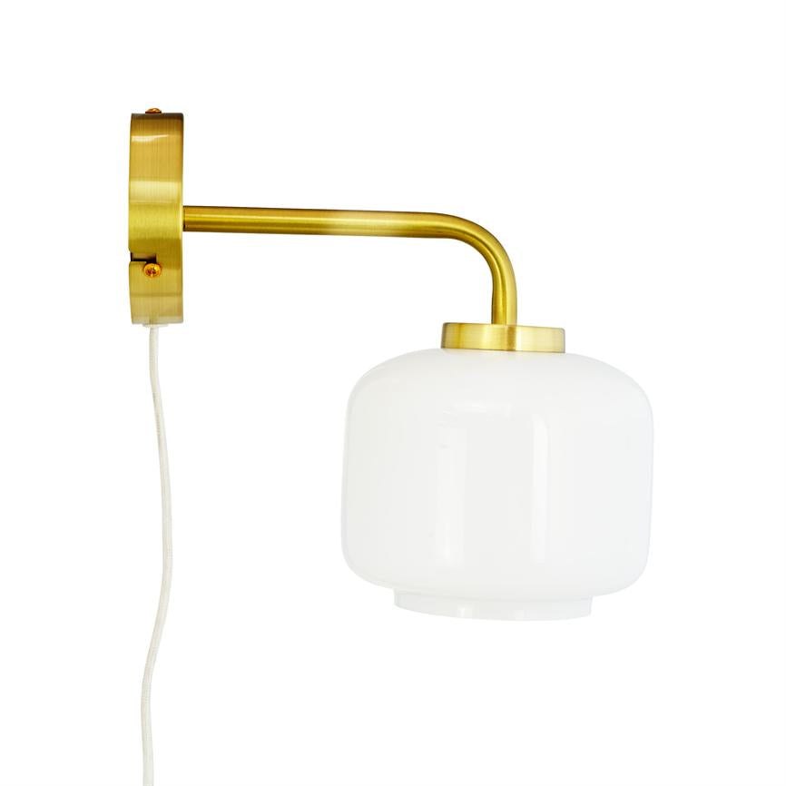 Væglampe hænger fra en buet, børstet messingarm; en rund opalhvid glasskærm vender nedad; lampen er monteret på en rund messingplade mod en hvid baggrund.