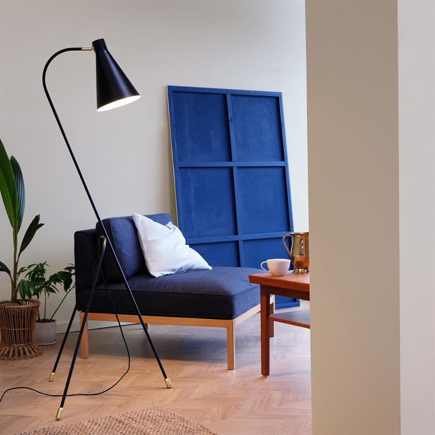 En mørkeblå loungestol står med en hvid pude; en sort gulvlampe lyser mod stolen, et blåt lærred læner sig mod væggen, omgivet af et træbord, planter og lyst trægulv.