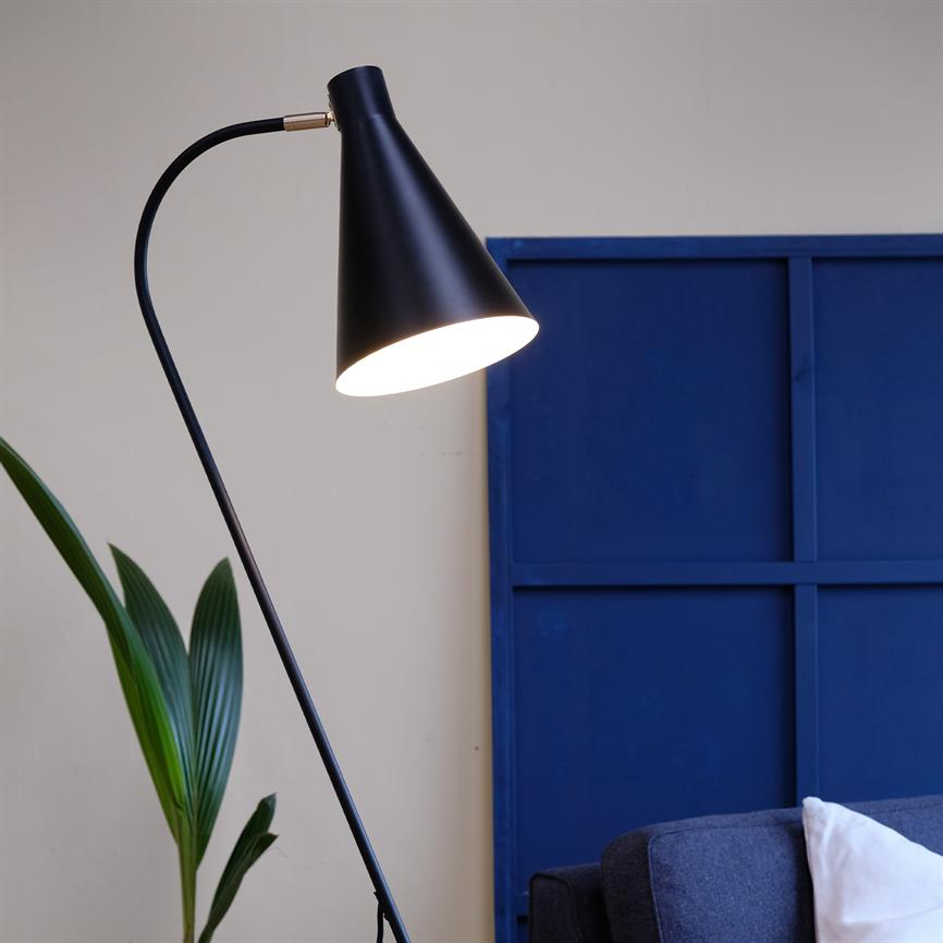 Sort, kegleformet gulvlampe lyser nedad ved siden af en mørkegrå sofa; står foran et stort blåt panel og ved en grøn stueplante i et lyst, neutralt rum.