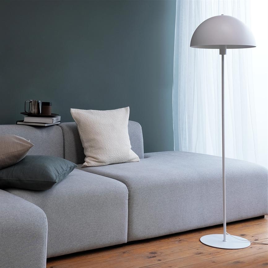 En lysegrå modulsofa er møbleret med puder og bøger; en hvid gulvlampe står ved et lyst, gardinklædt vindue i et roligt, mørkegrønt rum med trægulv.