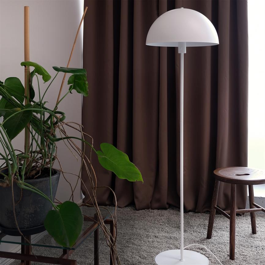 Hvid halvkugleformet gulvlampe står foran mørkebrune gardiner på et gråt tæppe, flankeret af en stor krukkeplante med lange lianer og en træskammel ved vinduet.