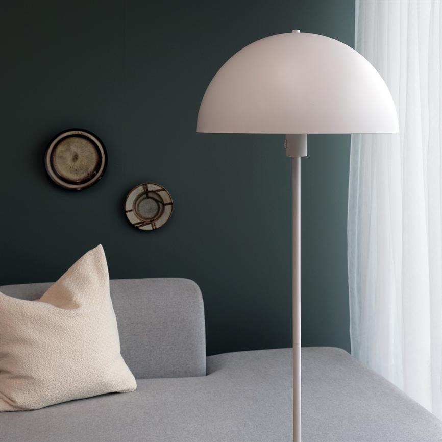 En hvid gulvlampe står ved siden af en grå sofa; en cremefarvet pude hviler på sofaen, en mørkegrøn væg med to dekorative tallerkener og hvide gardiner i baggrunden.